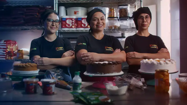 Três confeiteiras de braços cruzados atrás dos bolos